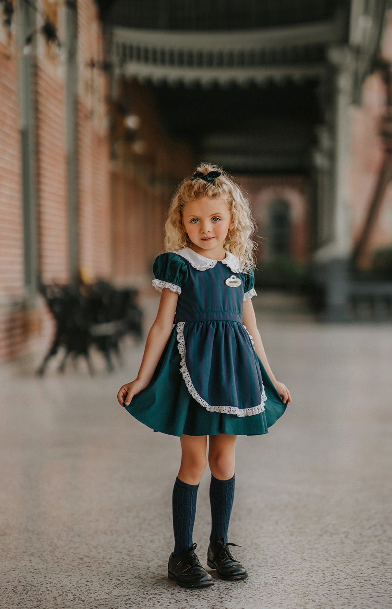 A young girl stands indoors wearing a dark teal green dress with white lace trim, a matching plaid blue and green apron, puff sleeves and a white collar.  The woven cotton is perfect for spooky season.