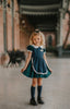 A young girl stands indoors wearing a dark teal green dress with white lace trim, a matching plaid blue and green apron, puff sleeves and a white collar.  The woven cotton is perfect for spooky season.