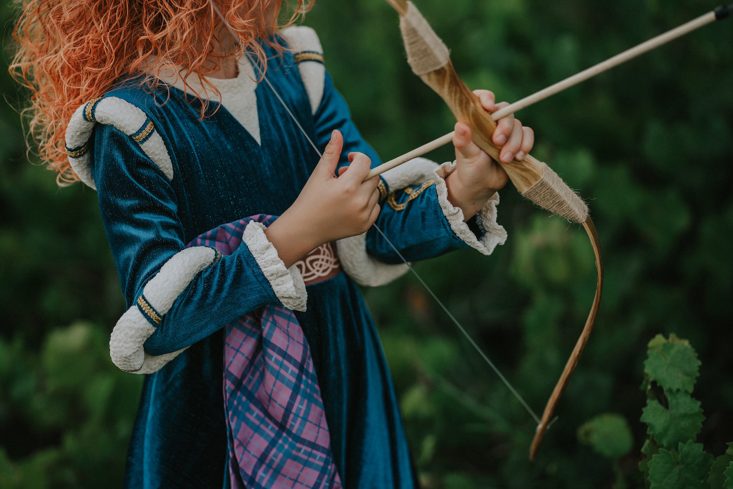 A young girl is wearing a long peacock blue green velvet gown.  The velvet has silver sparkle. The long sleeves have off white shoulder and elbow detail.  There is gold rope trim around the skirt.  A Celtic pattern band in tan has a purple and blue plaid sash with a gold button accent.  The base of the dress has a Celtic inspired pattern in shades of blue green. 