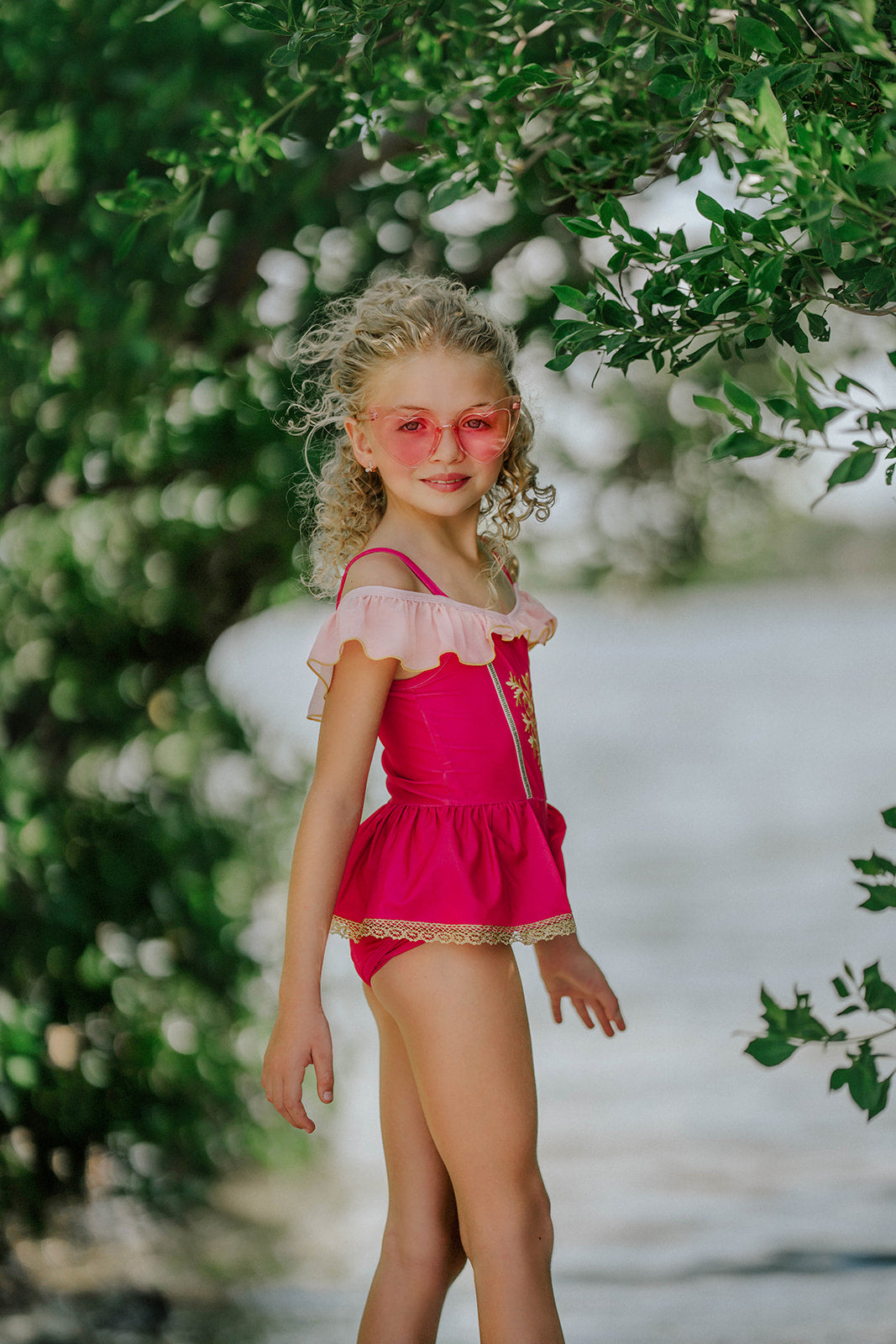 Girl wearing pink princess-inspired swimsuit with gold embroidery and ruffle off-shoulder detail, posing barefoot by the water

Girls royal-themed pink and gold one-piece swimsuit with peplum skirt, styled with ruffled straps and heart sunglasses

Princess bathing suit for girls in hot pink with gold lace trim and off-shoulder ruffles, perfect for summer dress-up swimwear