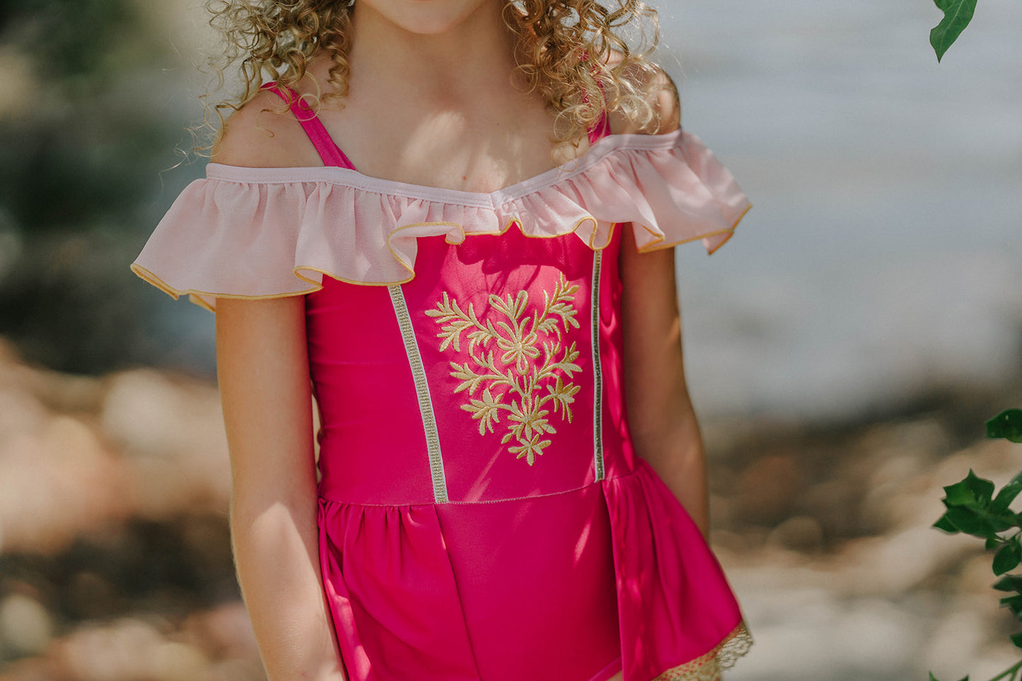 Girl wearing pink princess-inspired swimsuit with gold embroidery and ruffle off-shoulder detail, posing barefoot by the water

Girls royal-themed pink and gold one-piece swimsuit with peplum skirt, styled with ruffled straps and heart sunglasses

Princess bathing suit for girls in hot pink with gold lace trim and off-shoulder ruffles, perfect for summer dress-up swimwear
