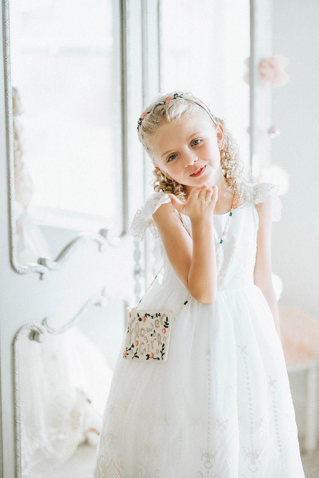 Child’s white flower girl gown with lace sleeves and bow back, styled with a beaded gold and pink floral purse for weddings