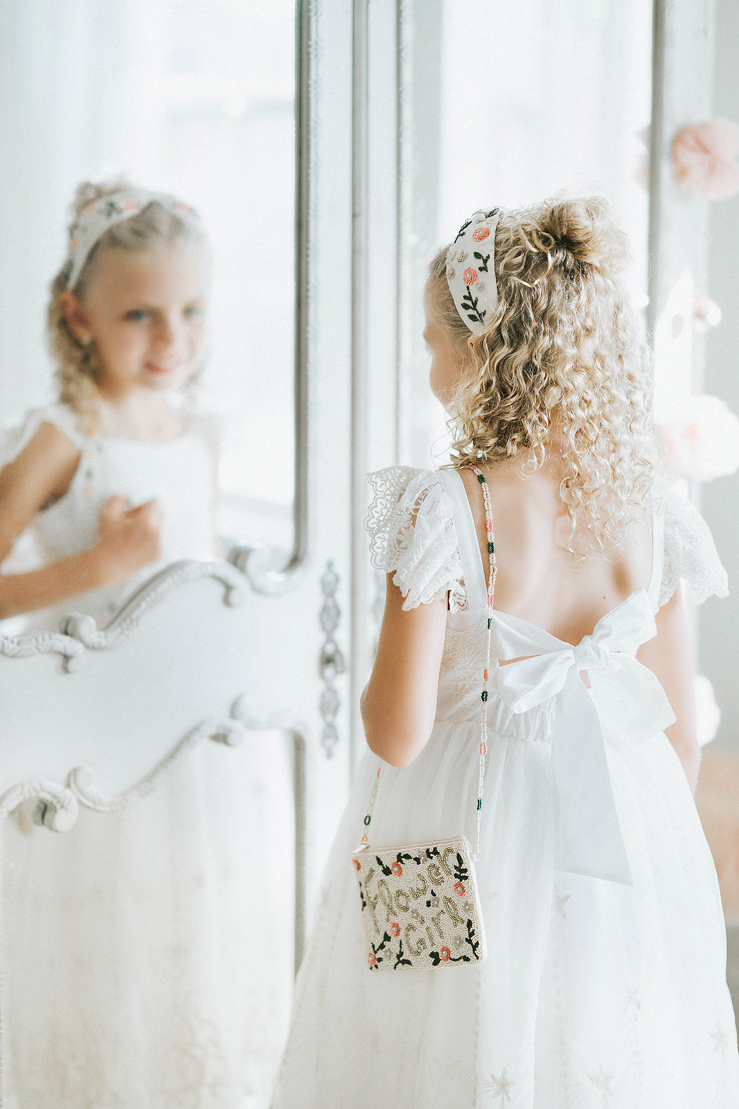 Elegant flower girl in white lace dress standing by mirror, wearing a vintage-style beaded purse that reads “Flower Girl”