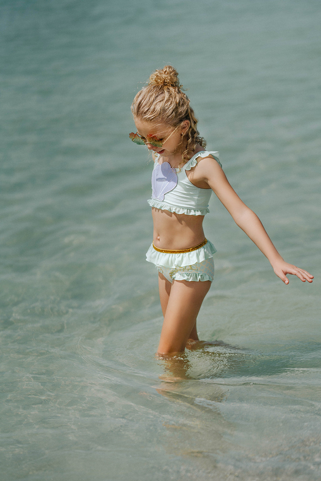 Girl wearing mint green mermaid swimsuit with purple seashell top and gold waistband, standing at the shoreline in fun sunglasses

Mermaid-themed ruffle bikini for girls with shell applique and scale print bottoms, styled for beach photos and play

Child in seafoam green two-piece swimsuit with frilly straps and shell detail, posing on sandy beach in sunshine