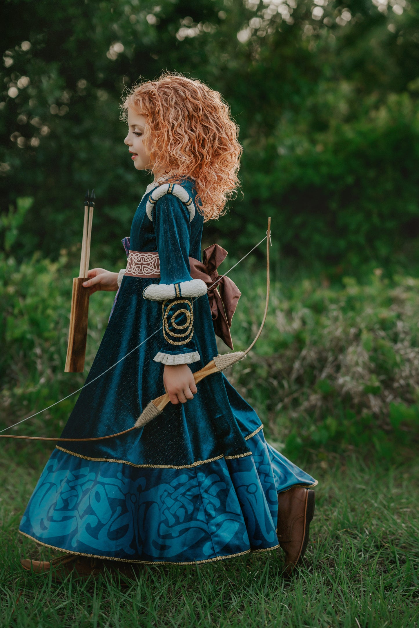 A young girl is wearing a long peacock blue green velvet gown.  The velvet has silver sparkle. The long sleeves have off white shoulder and elbow detail.  There is gold rope trim around the skirt.  A Celtic pattern band in tan has a purple and blue plaid sash with a gold button accent.  The base of the dress has a Celtic inspired pattern in shades of blue green. 