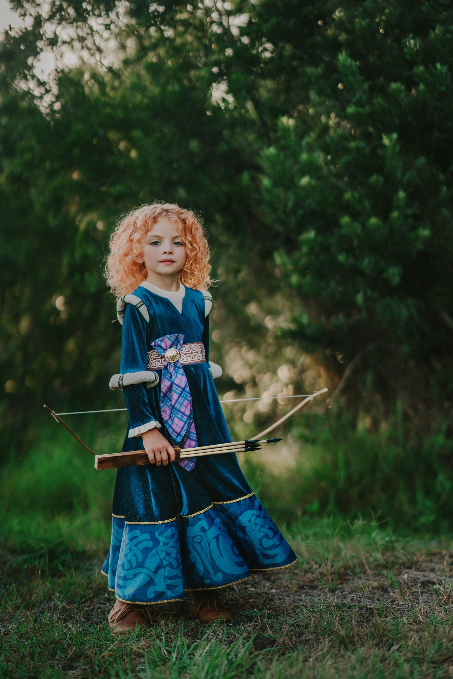 A young girl is wearing a long peacock blue green velvet gown.  The velvet has silver sparkle. The long sleeves have off white shoulder and elbow detail.  There is gold rope trim around the skirt.  A Celtic pattern band in tan has a purple and blue plaid sash with a gold button accent.  The base of the dress has a Celtic inspired pattern in shades of blue green. 