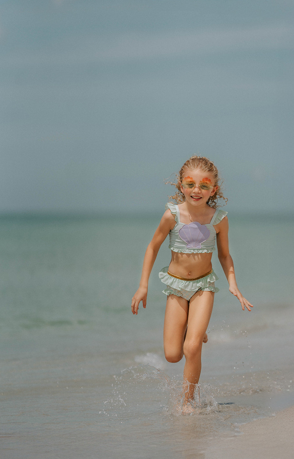 Child in seafoam green two-piece swimsuit with frilly straps and shell detail, posing on sandy beach in sunshine

Girls pastel mermaid swimsuit with purple shell top and ruffled bottoms, styled with gold accents and seashell sunglasses

Toddler to tween girl in ruffled shell-themed mermaid swimsuit with sparkle waistband, perfect for summer vacation