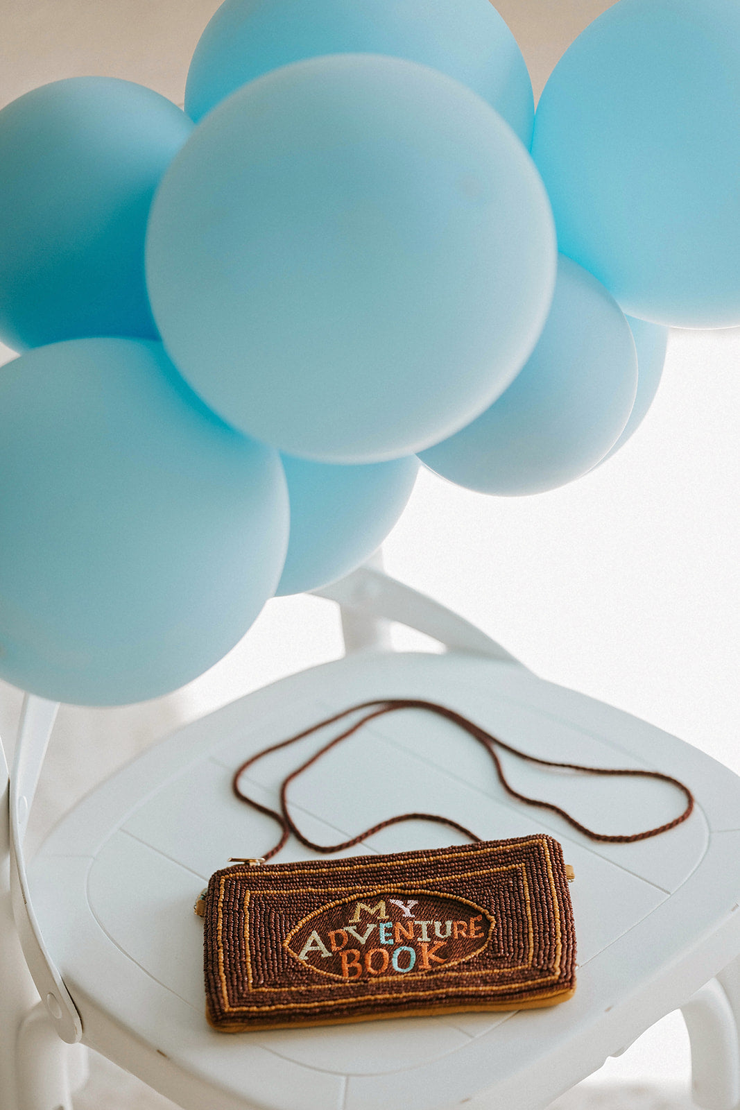 Brown beaded clutch purse with My Adventure Book cover design and multicolor lettering, styled with light blue balloons for an Up birthday party or travel-themed photoshoot.

Pixar Up–inspired beaded handbag with whimsical book cover styling, ideal for gifting to Disney lovers or little explorers heading to Disneyland or a balloon-themed party.

Adventurous crossbody bag for girls or women styled like the Up movie scrapbook, featuring intricate seed bead design and rope-style strap, photographed with pastel