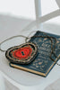 Beaded heart purse with red and black design, keyhole detail, and intricate gold stitching, styled with a white dress and blue tartan scarf for a fairytale holiday accessory look.

Gothic-inspired red heart handbag with keyhole motif and beadwork, worn by girl in winter outfit, perfect for Valentine’s Day, fairytale fashion, or themed photoshoots.

Close-up of a heart-shaped beaded purse with ornate swirls, pearl accents, and a romantic keyhole center, styled for girls or women who love storybook and fantas