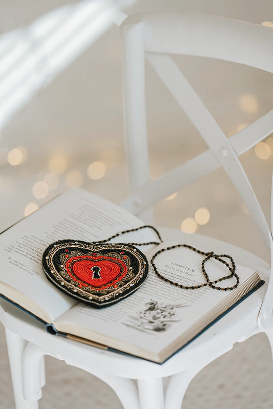 Beaded heart purse with red and black design, keyhole detail, and intricate gold stitching, styled with a white dress and blue tartan scarf for a fairytale holiday accessory look.

Gothic-inspired red heart handbag with keyhole motif and beadwork, worn by girl in winter outfit, perfect for Valentine’s Day, fairytale fashion, or themed photoshoots.

Close-up of a heart-shaped beaded purse with ornate swirls, pearl accents, and a romantic keyhole center, styled for girls or women who love storybook and fantas