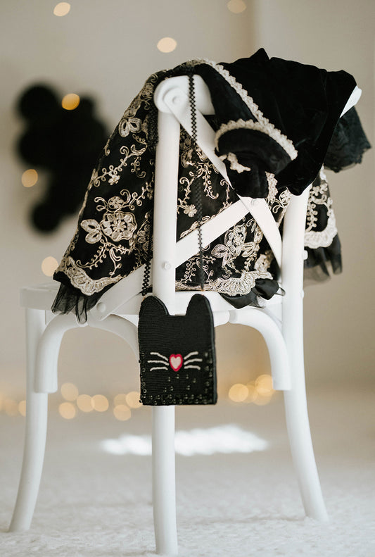 Black beaded cat purse with heart-shaped nose and whiskers, styled with lace-trimmed gothic dress on a white chair for Halloween trick-or-treat or costume party accessory.

Whimsical black cat crossbody bag for girls with beaded design, heart nose, and whiskers, styled next to embroidered Halloween dress for spooky season outfit inspiration.

Close-up of handmade cat-shaped purse featuring sparkly black seed beads and pink heart nose, hanging from white chair for Halloween, gothic, or dress-up styling.