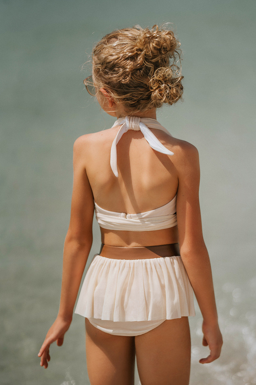 Boho-inspired cream girls swimsuit with wrap detail and side draping, styled for beach photos or summer vacation

Toddler to tween girl in goddess-style cream bathing suit with halter neckline and high-waisted bottoms, standing on the shore

Girls neutral swimwear set featuring wrap halter top and soft mesh skirt, perfect for boho beachwear or summer shoots