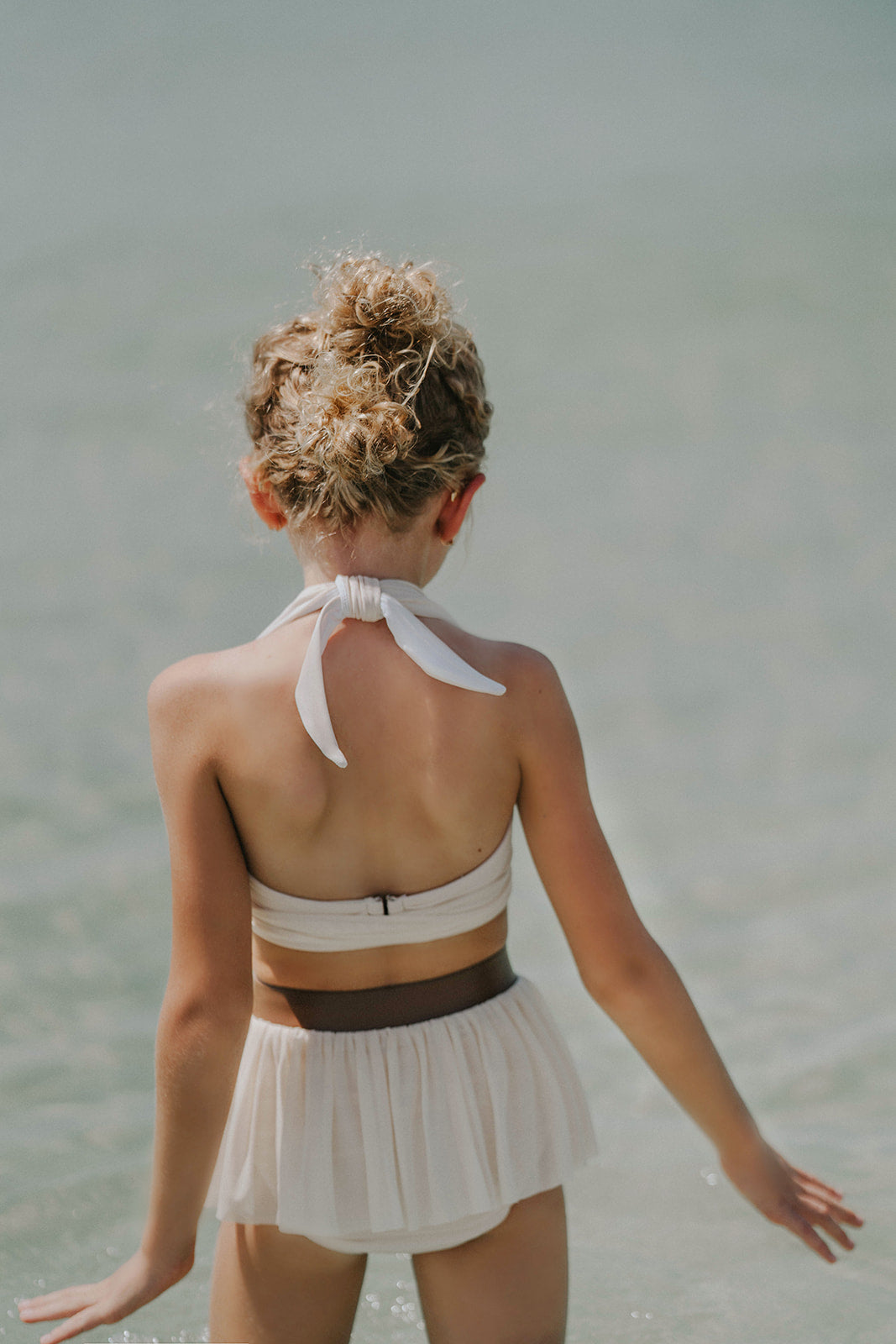 Toddler to tween girl in goddess-style cream bathing suit with halter neckline and high-waisted bottoms, standing on the shore

Girls neutral swimwear set featuring wrap halter top and soft mesh skirt, perfect for boho beachwear or summer shoots

Child in cream-colored two-piece swimsuit with wrap top and skirted bottom, posing barefoot in the sand on a sunny beach day