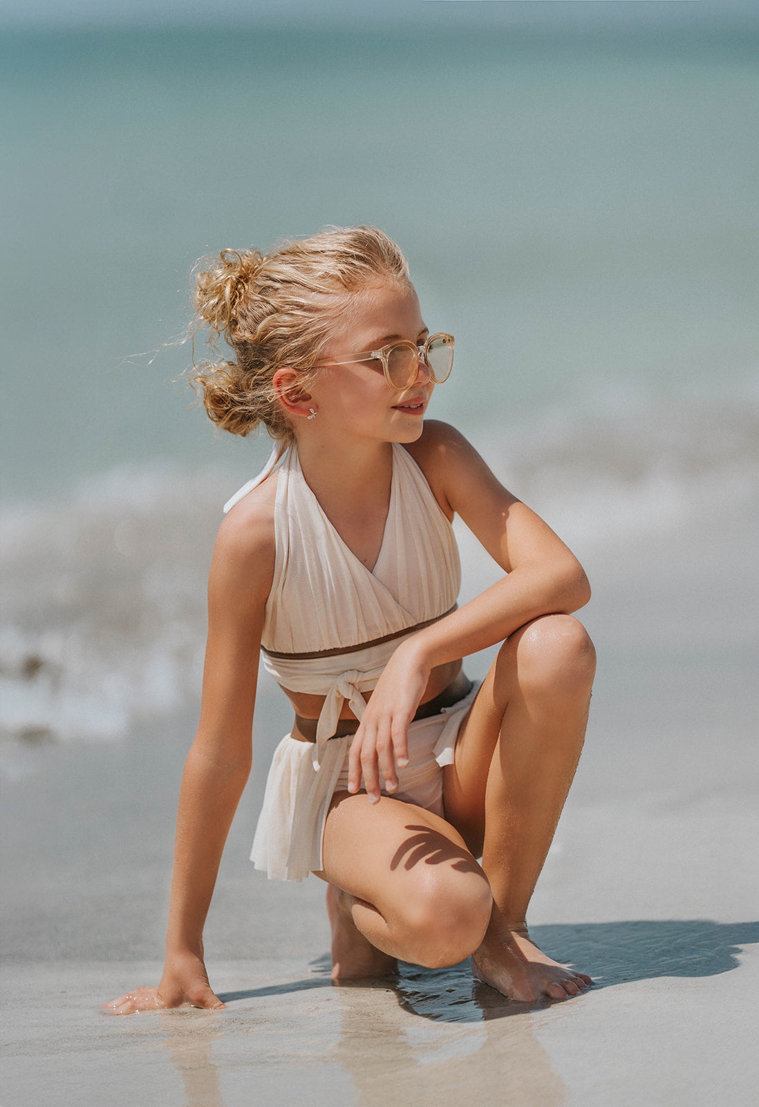 Boho-inspired cream girls swimsuit with wrap detail and side draping, styled for beach photos or summer vacation

Toddler to tween girl in goddess-style cream bathing suit with halter neckline and high-waisted bottoms, standing on the shore

Girls neutral swimwear set featuring wrap halter top and soft mesh skirt, perfect for boho beachwear or summer shoots

Child in cream-colored two-piece swimsuit with wrap top and skirted bottom, posing barefoot in the sand on a sunny beach day