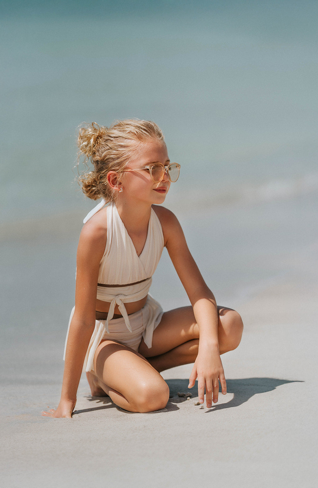 Boho-inspired cream girls swimsuit with wrap detail and side draping, styled for beach photos or summer vacation

Toddler to tween girl in goddess-style cream bathing suit with halter neckline and high-waisted bottoms, standing on the shore

Girls neutral swimwear set featuring wrap halter top and soft mesh skirt, perfect for boho beachwear or summer shoots

Child in cream-colored two-piece swimsuit with wrap top and skirted bottom, posing barefoot in the sand on a sunny beach day
