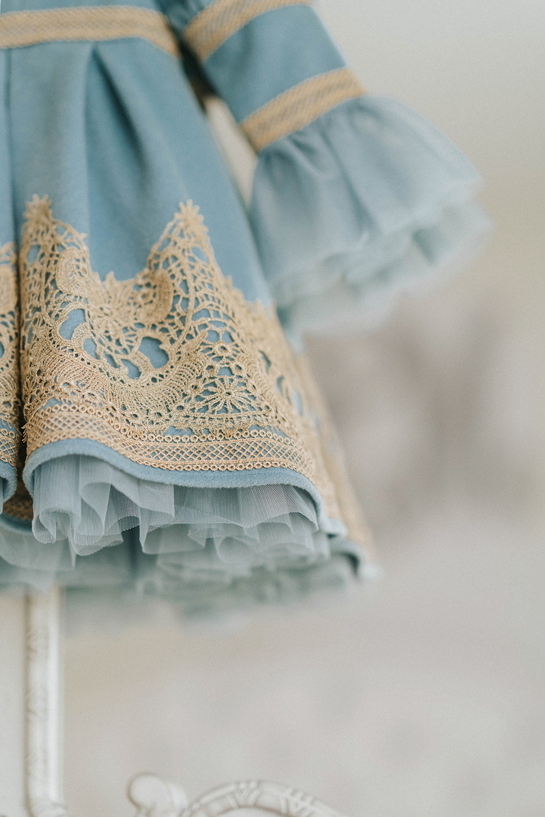 Elegant heirloom girls dress in dusty blue with gold lace trim and tulle layers, hanging on ornate vintage-style mirror

Long sleeve blue and gold embroidered holiday dress for girls, featuring pearl buttons and layered tulle hem

Girls vintage-style formal gown in soft blue with gold lace and flounce sleeves, ideal for portraits or weddings

Classic blue heirloom dress for toddlers and girls with ornate embroidery and pearl detailing, styled for a timeless photo shoot