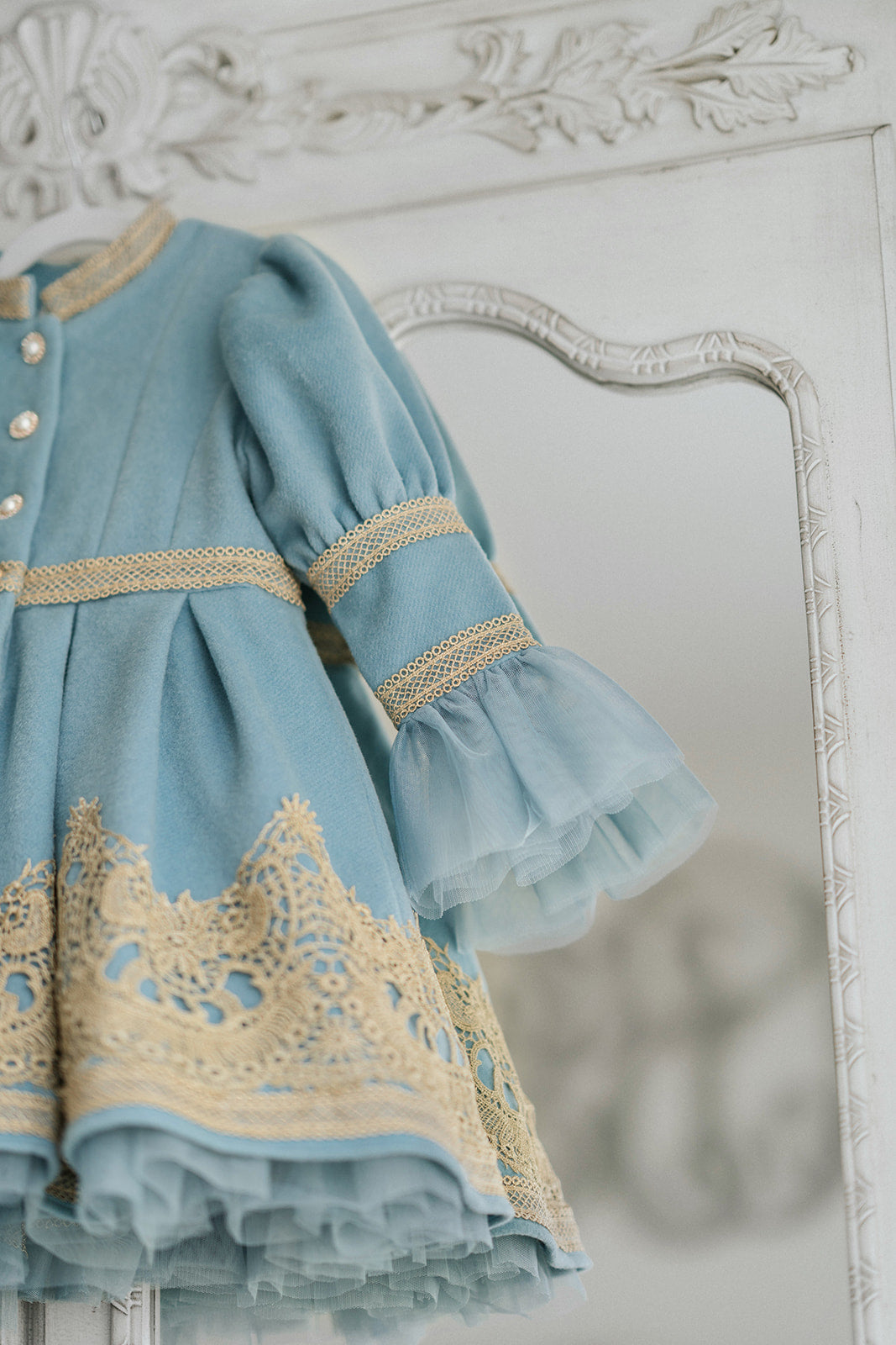 Elegant heirloom girls dress in dusty blue with gold lace trim and tulle layers, hanging on ornate vintage-style mirror

Long sleeve blue and gold embroidered holiday dress for girls, featuring pearl buttons and layered tulle hem

Girls vintage-style formal gown in soft blue with gold lace and flounce sleeves, ideal for portraits or weddings

Classic blue heirloom dress for toddlers and girls with ornate embroidery and pearl detailing, styled for a timeless photo shoot