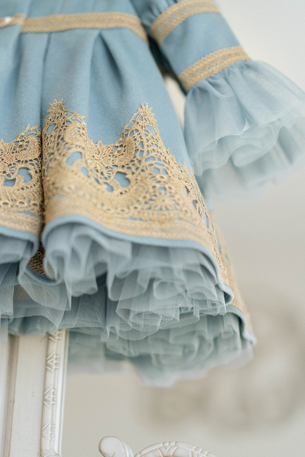 Elegant heirloom girls dress in dusty blue with gold lace trim and tulle layers, hanging on ornate vintage-style mirror

Long sleeve blue and gold embroidered holiday dress for girls, featuring pearl buttons and layered tulle hem

Girls vintage-style formal gown in soft blue with gold lace and flounce sleeves, ideal for portraits or weddings

Classic blue heirloom dress for toddlers and girls with ornate embroidery and pearl detailing, styled for a timeless photo shoot