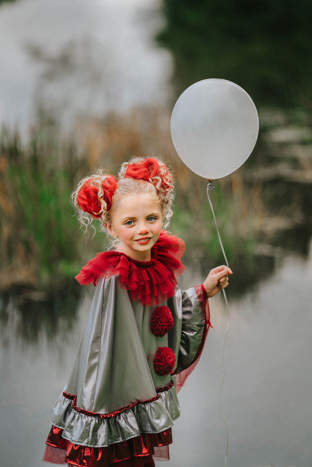 Dancing clown costume for girls in shiny silver and red with dramatic pom-poms and a layered ruffle neckline, perfect for dress-up

Young girl in vintage circus-inspired clown dress holding a balloon, wearing metallic cape with red trim and curly hair buns

Theatrical clown costume for kids with metallic silver fabric and bold red accents, ideal for Halloween or costume party photos