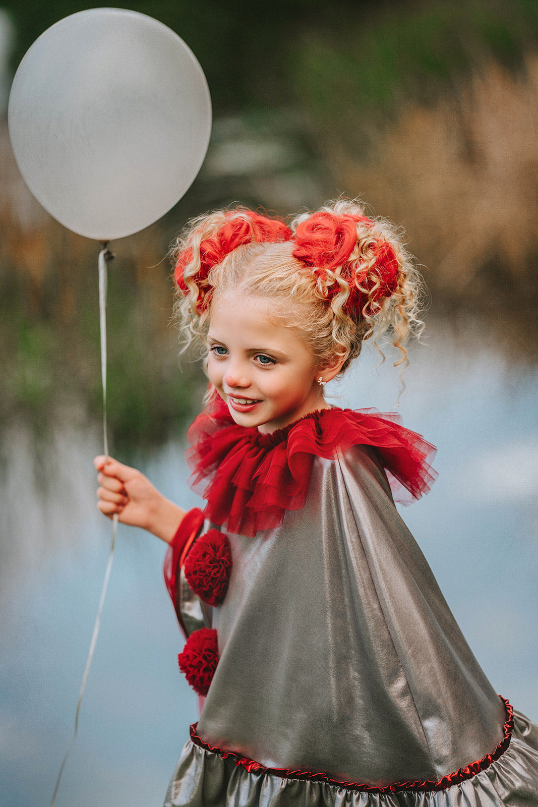 Dancing clown costume for girls in shiny silver and red with dramatic pom-poms and a layered ruffle neckline, perfect for dress-up

Young girl in vintage circus-inspired clown dress holding a balloon, wearing metallic cape with red trim and curly hair buns

Theatrical clown costume for kids with metallic silver fabric and bold red accents, ideal for Halloween or costume party photos