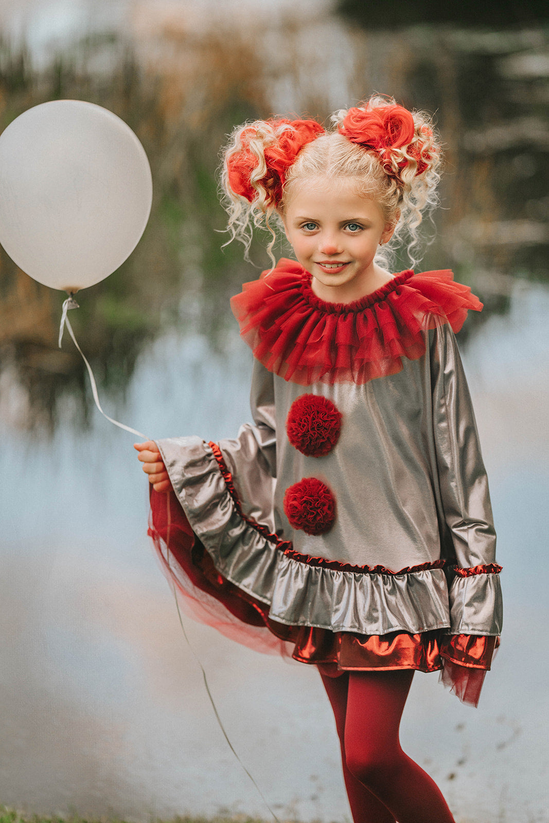 Girl wearing silver and red clown costume with ruffled collar and pom-poms, holding a white balloon near a pond in whimsical setting

Child dressed in metallic clown cape with red tights and tulle ruffle collar, styled for Halloween or circus-themed photoshoot

Dancing clown costume for girls in shiny silver and red with dramatic pom-poms and a layered ruffle neckline, perfect for dress-up