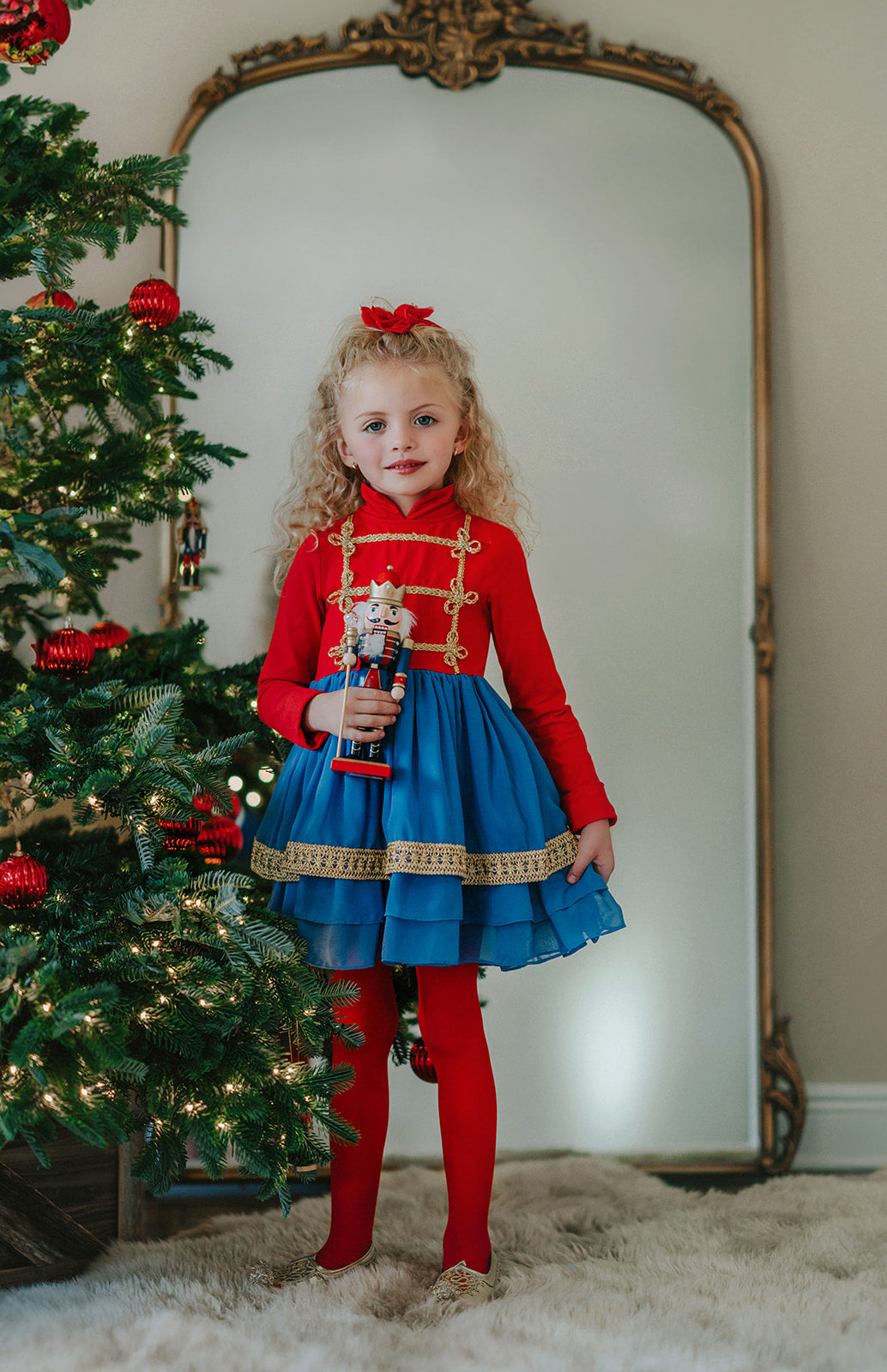 A child standing in front of a decorated Christmas tree, wearing a red and blue twirl dress with gold braiding, holding a toy, in a room with a mirror behind her.