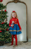 A child standing in front of a decorated Christmas tree, wearing a red and blue twirl dress with gold braiding, holding a toy, in a room with a mirror behind her.