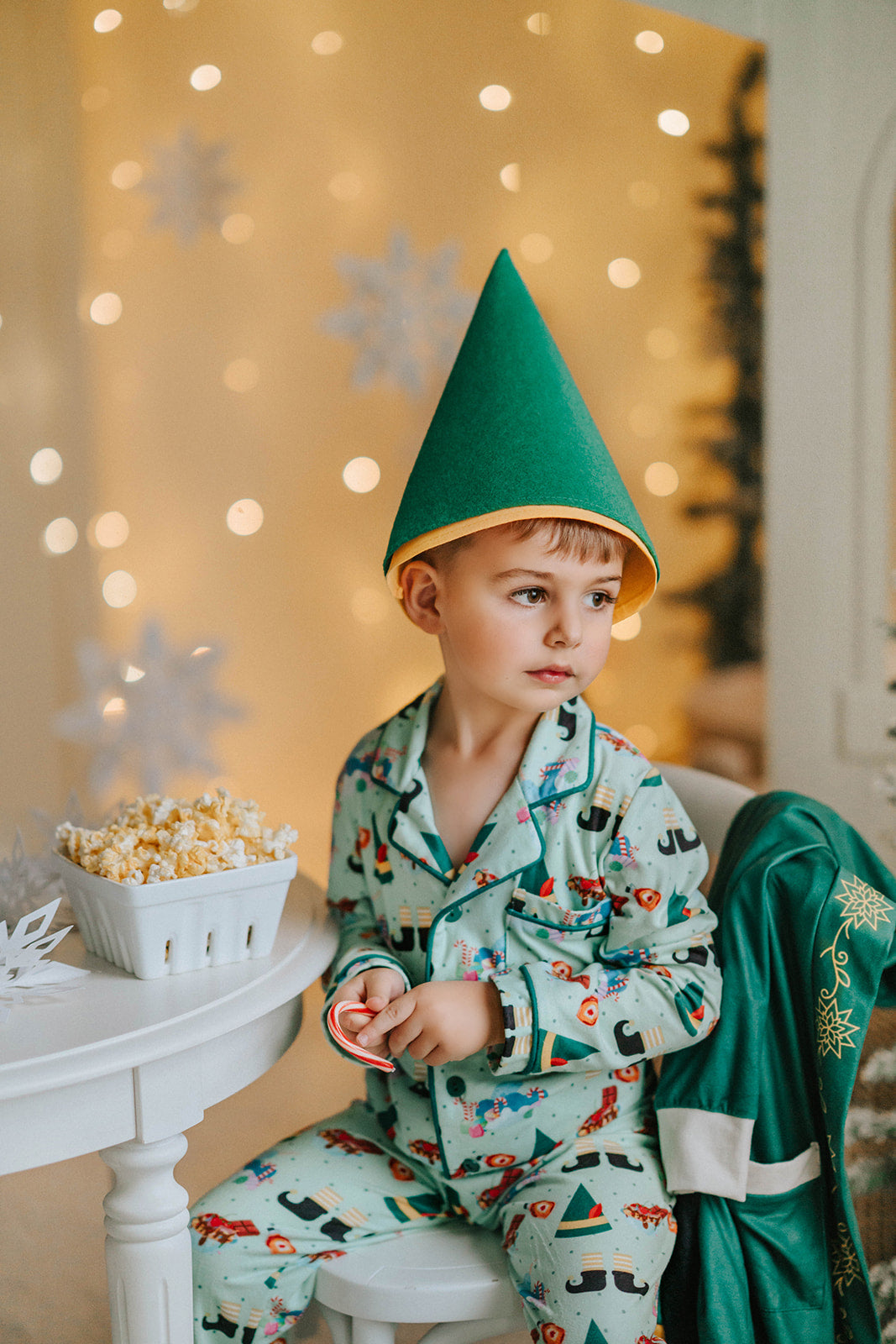 Toddler boy in festive Christmas pajamas and green elf hat standing near Christmas trees and lights, holding ribbon

Young boy in holiday pajama set with elf hat, surrounded by snow-covered trees and popcorn, ready for a Christmas photo

Child dressed in green Christmas pajamas with matching elf cone hat, standing in a winter-themed holiday setup