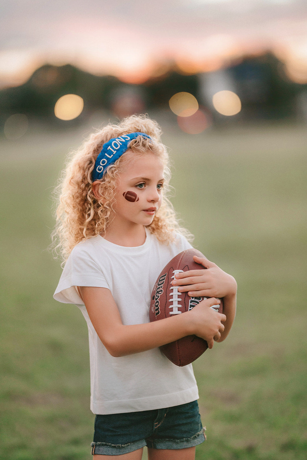 Girl wearing blue “Go Lions” headband holding football on a field at sunset, dressed in white tee and denim shorts

Child wearing sparkly blue team spirit headband that says “Go Lions,” styled with football face sticker and sporty outfit

Little girl showing team pride in “Go Lions” beaded headband while holding football during golden hour on the field

Girls football game day outfit with blue “Go Lions” headband and pigskin, perfect for team spirit wear or cheerleaders