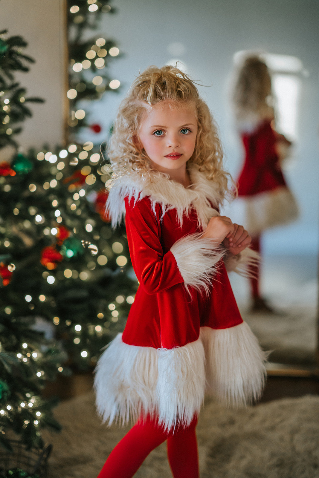 A young girl stands at a Christmas tree in the Martha Stanta Dress.  This dress is a true red soft crushed velvet.  The collar, front, cuffs and hem are lined with a white fluffy fur.  There is a black belt with silver buckle at the waist. 