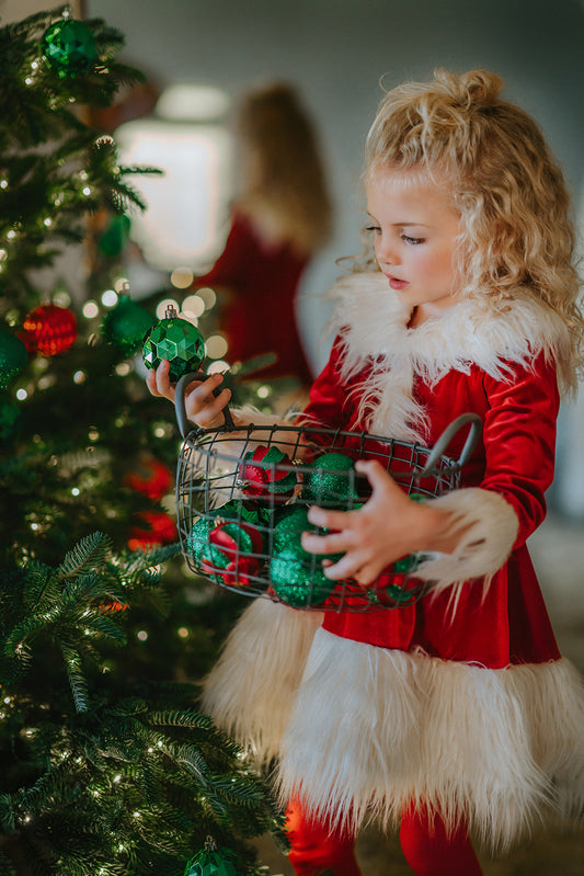 A young girl stands at a Christmas tree in the Martha Stanta Dress.  This dress is a true red soft crushed velvet.  The collar, front, cuffs and hem are lined with a white fluffy fur.  There is a black belt with silver buckle at the waist. 