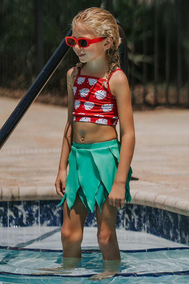 Girl wearing red monstera crop top and green leaf skirt swimsuit standing by the pool in tropical-themed two-piece swimwear

Toddler girl in Hawaiian two-piece swimsuit with red monstera print top and layered green leaf skirt at the poolside

Child in tropical swim outfit with red printed crop top and green leaf petal skirt, styled for luau or summer vacation