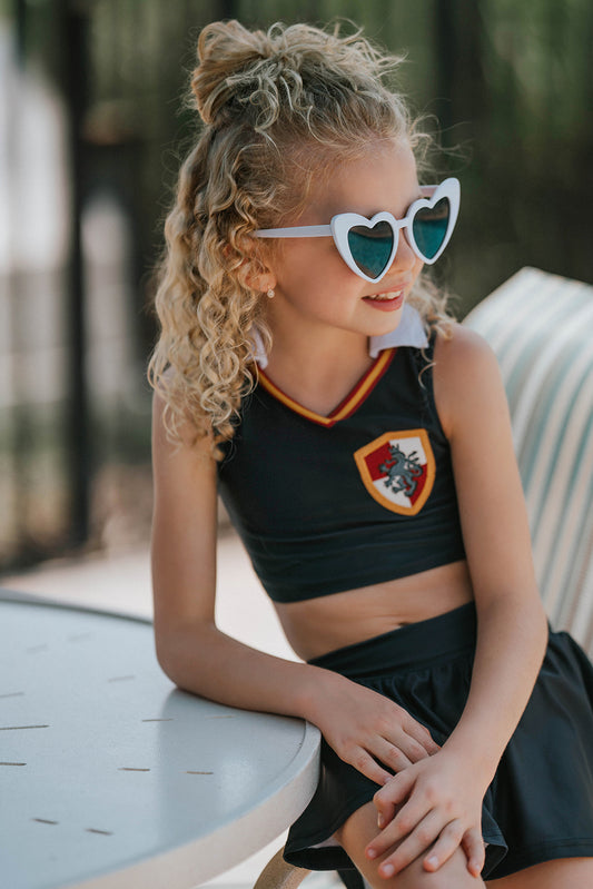 Blonde young girl standing on a pale-sand beach wearing a black two-piece athletic swim set—cropped tank top and skirted bottoms—with a subtle wand-and-star wizard emblem on the chest. The scene shows gentle turquoise waves in the background
