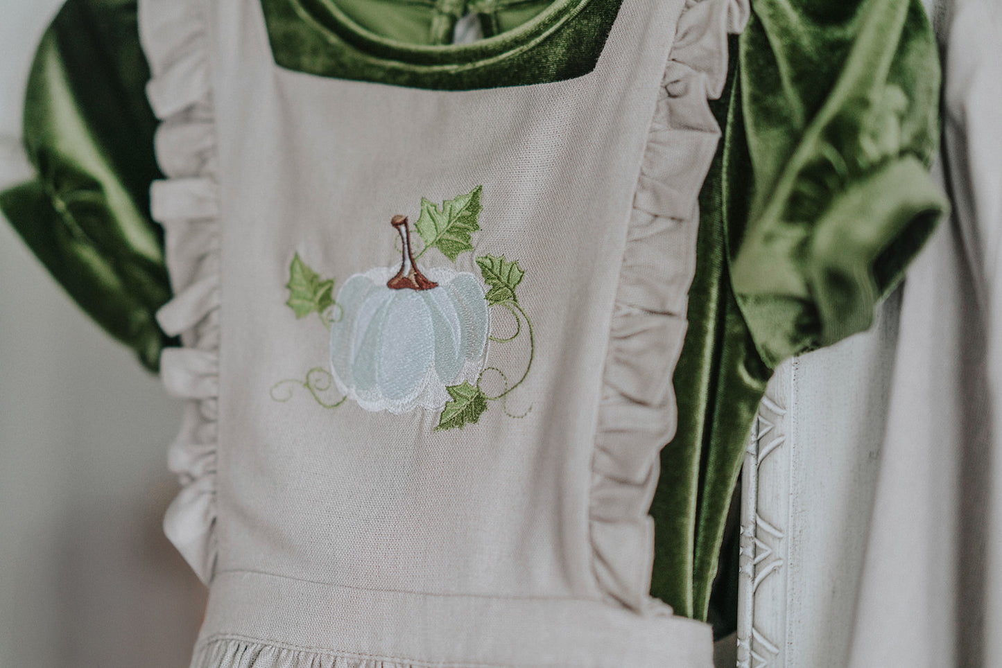 Girls green velvet dress with linen apron featuring embroidered pale blue pumpkin and ruffle trim, perfect for fall photos or Thanksgiving

Heirloom-style green velvet holiday dress with linen pinafore and pumpkin embroidery, styled for autumn or fairytale dress-up

Fall girls outfit with green velvet dress and embroidered pumpkin apron, hanging on vintage white carved mirror

Toddler to tween Thanksgiving dress with velvet base, apron pockets, and embroidered pumpkin design, ideal for family photos