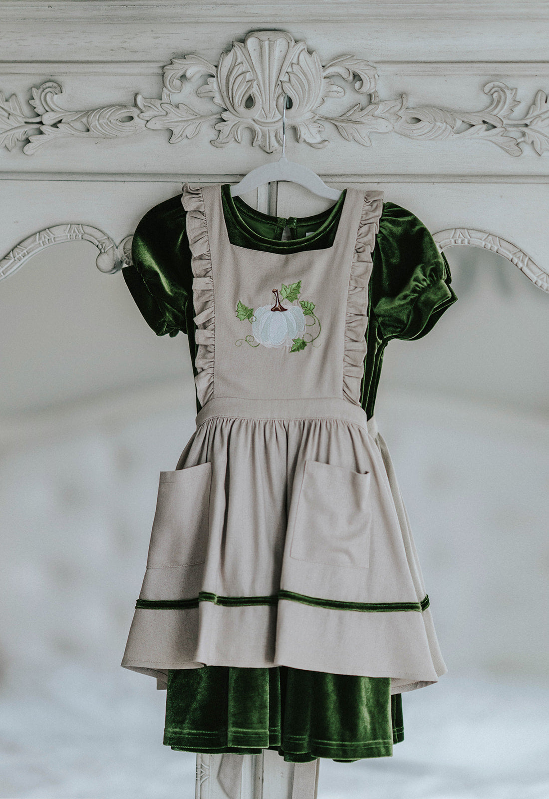 Girls green velvet dress with linen apron featuring embroidered pale blue pumpkin and ruffle trim, perfect for fall photos or Thanksgiving

Heirloom-style green velvet holiday dress with linen pinafore and pumpkin embroidery, styled for autumn or fairytale dress-up

Fall girls outfit with green velvet dress and embroidered pumpkin apron, hanging on vintage white carved mirror

Toddler to tween Thanksgiving dress with velvet base, apron pockets, and embroidered pumpkin design, ideal for family photos