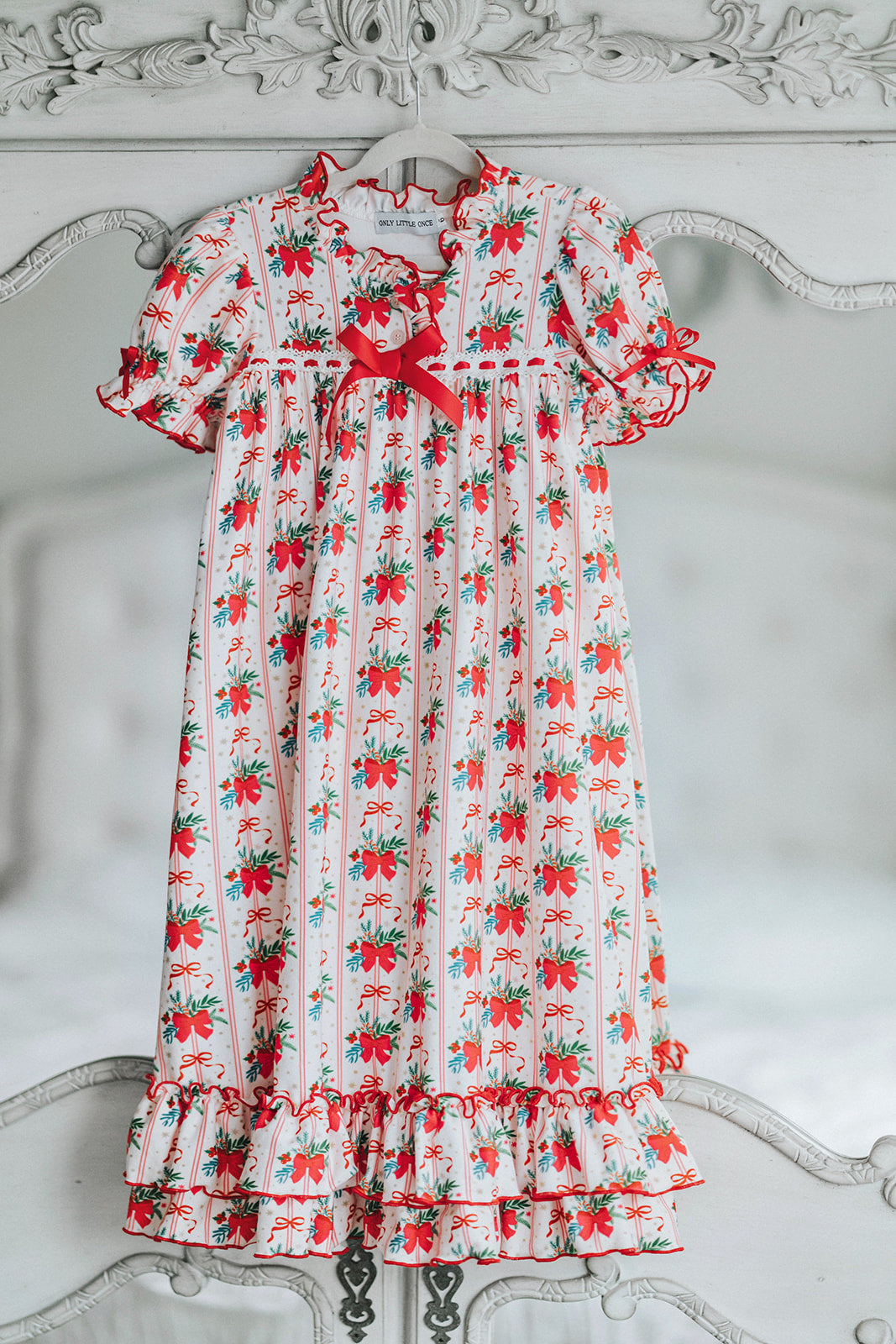 Vintage-style girls Christmas nightgown with red bows, holly print, ruffle trim, and satin bow, hanging on a carved white mirror

Classic holiday pajama dress for girls with red and green holly print, ruffle hem, and red satin bow at the neckline

Girls long sleeve Christmas nightgown with festive red bow and holly pattern, styled for holiday photos or heirloom gifting

Traditional girls holiday nightgown in white with red and green floral print, ruffle accents, and lace trim