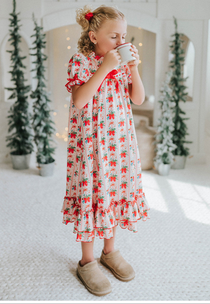 Vintage-style girls Christmas nightgown with red bows, holly print, ruffle trim, and satin bow, hanging on a carved white mirror

Classic holiday pajama dress for girls with red and green holly print, ruffle hem, and red satin bow at the neckline

Girls long sleeve Christmas nightgown with festive red bow and holly pattern, styled for holiday photos or heirloom gifting

Traditional girls holiday nightgown in white with red and green floral print, ruffle accents, and lace trim
