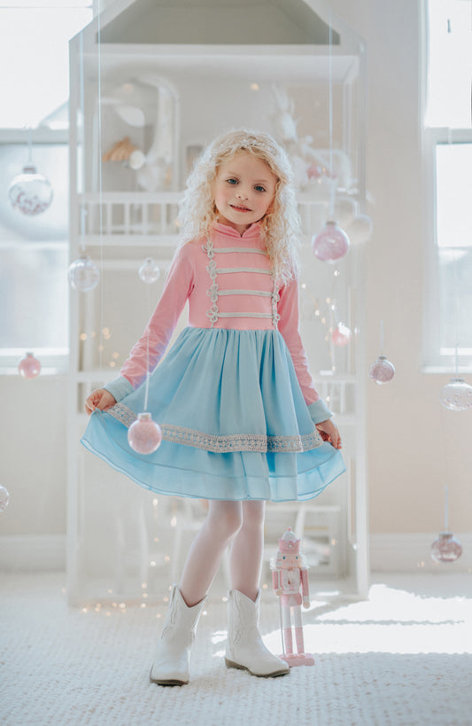 A young girl in a pastel toy soldier dress with a mandarin collar, intricate chest details, and a long skirt, standing in a room with Christmas-themed decorations.