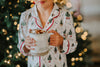 A young boy is wearing a traditional 2 piece lounge set.  The off white pjs have little christmas trees and christmas tree gnomes printed on it.  There is red piping on the top. 