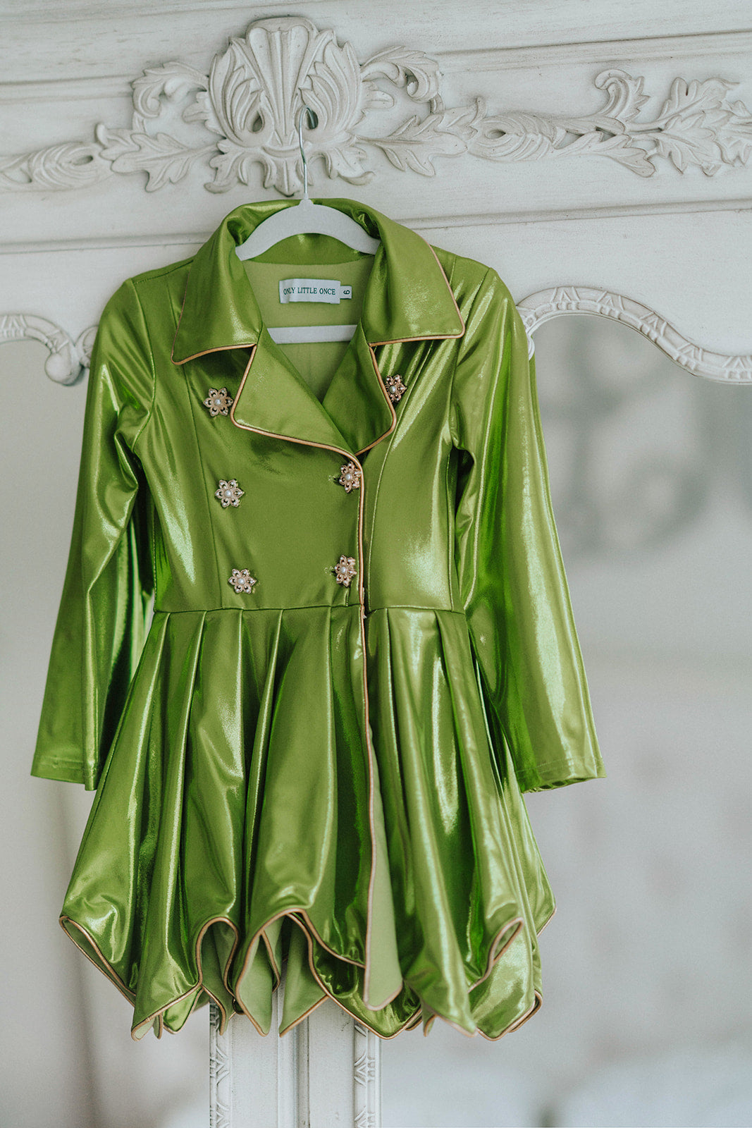 Girls metallic green coat dress with jeweled buttons and gold trim, displayed on mannequin against white brick wall

Shiny green holiday dress for girls with flared skirt, lapel collar, and crystal floral buttons, perfect for Christmas or pageants

Tinkerbell-inspired metallic green coat-style dress for girls, featuring pleated skirt and jeweled embellishments

Emerald green girls dress with long sleeves and coat silhouette, styled with gold piping and crystal button accents