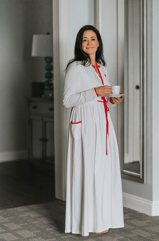 Woman wearing long white holiday nightgown with red trim, tie waist, and buttons, holding a teacup and smiling in a cozy morning setting, perfect for Christmas sleepwear.

White women's nightgown with red piping and buttons, styled with pockets and a tie waist, perfect for winter lounging, Christmas morning, or cozy holiday gift giving.

Adult holiday sleepwear for women featuring red contrast trim, full length, and soft knit fabric, styled with coffee cup in a warm, elegant home setting.
