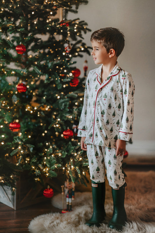 A young boy is wearing a traditional 2 piece lounge set.  The off white pjs have little christmas trees and christmas tree gnomes printed on it.  There is red piping on the top. 