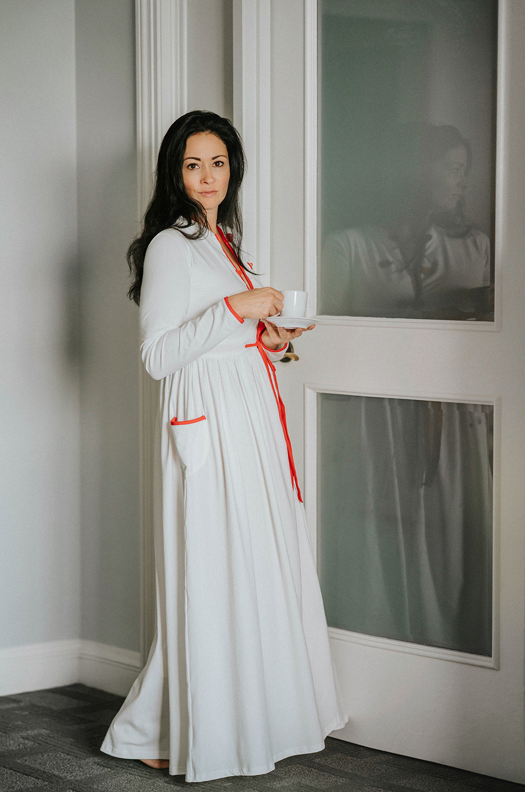 Woman wearing long white holiday nightgown with red trim, tie waist, and buttons, holding a teacup and smiling in a cozy morning setting, perfect for Christmas sleepwear.

White women's nightgown with red piping and buttons, styled with pockets and a tie waist, perfect for winter lounging, Christmas morning, or cozy holiday gift giving.

Adult holiday sleepwear for women featuring red contrast trim, full length, and soft knit fabric, styled with coffee cup in a warm, elegant home setting.