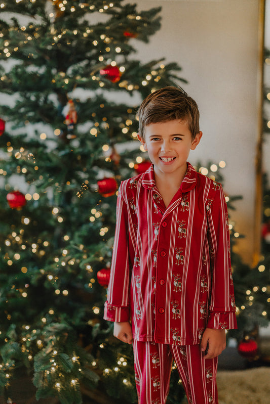 A young boy wearing a Unisex Reindeer Stripe Lounge set made from buttery soft fabric stands smiling in front of a decorated Christmas tree adorned with lights and red ornaments.