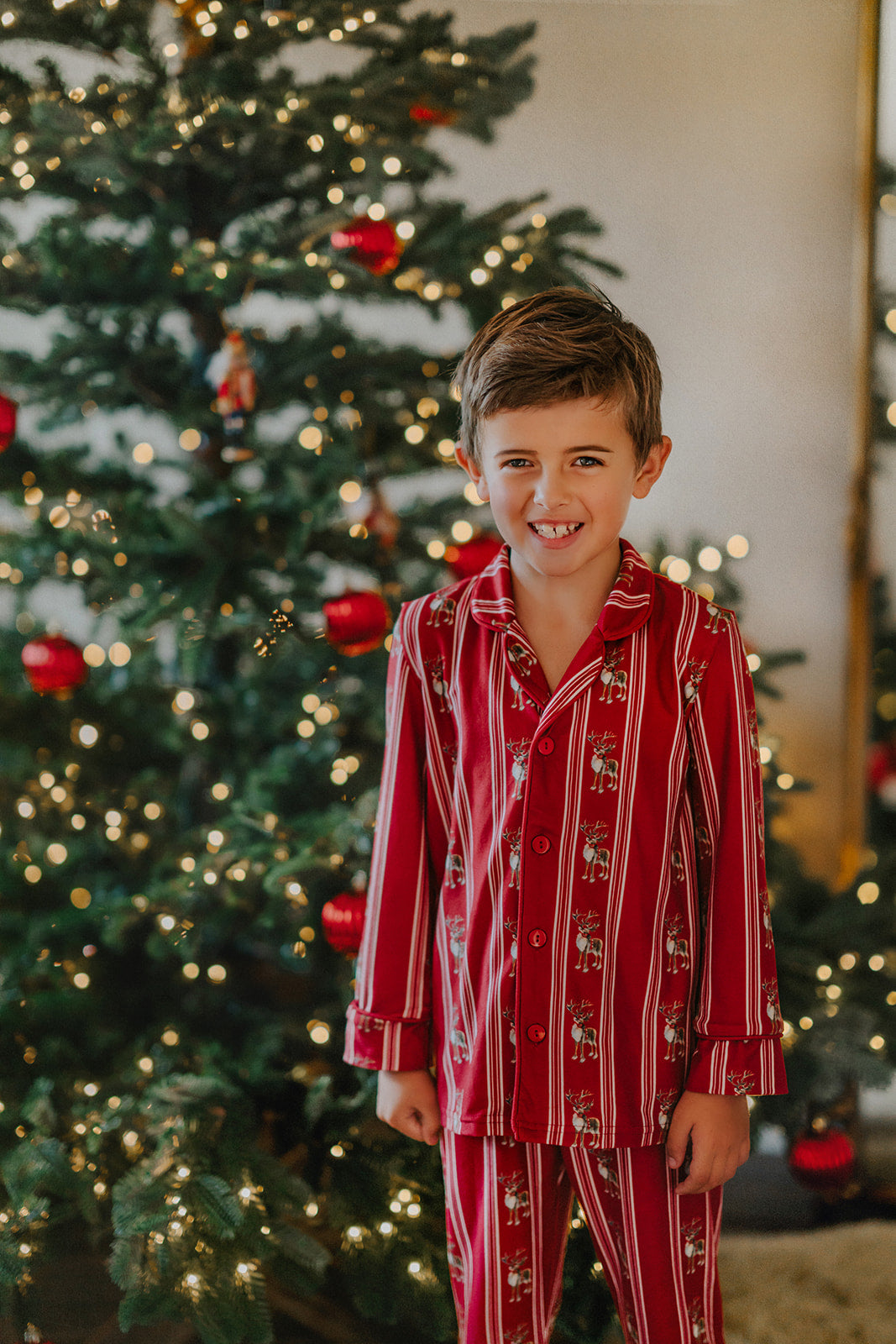 A young boy wearing a Unisex Reindeer Stripe Lounge set made from buttery soft fabric stands smiling in front of a Christmas tree. The 2 piece classic pajama set has reindeer and vertical cream colored stripes. 