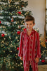 A young boy wearing a Unisex Reindeer Stripe Lounge set made from buttery soft fabric stands smiling in front of a Christmas tree. The 2 piece classic pajama set has reindeer and vertical cream colored stripes. 