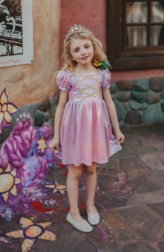 A young girls is wearing a metallic Rapunzel Twirl dress. The pink and purple knee length dress has a criss cross gold ribbon across the bodice. The ballroom puff sleeves add elegance.