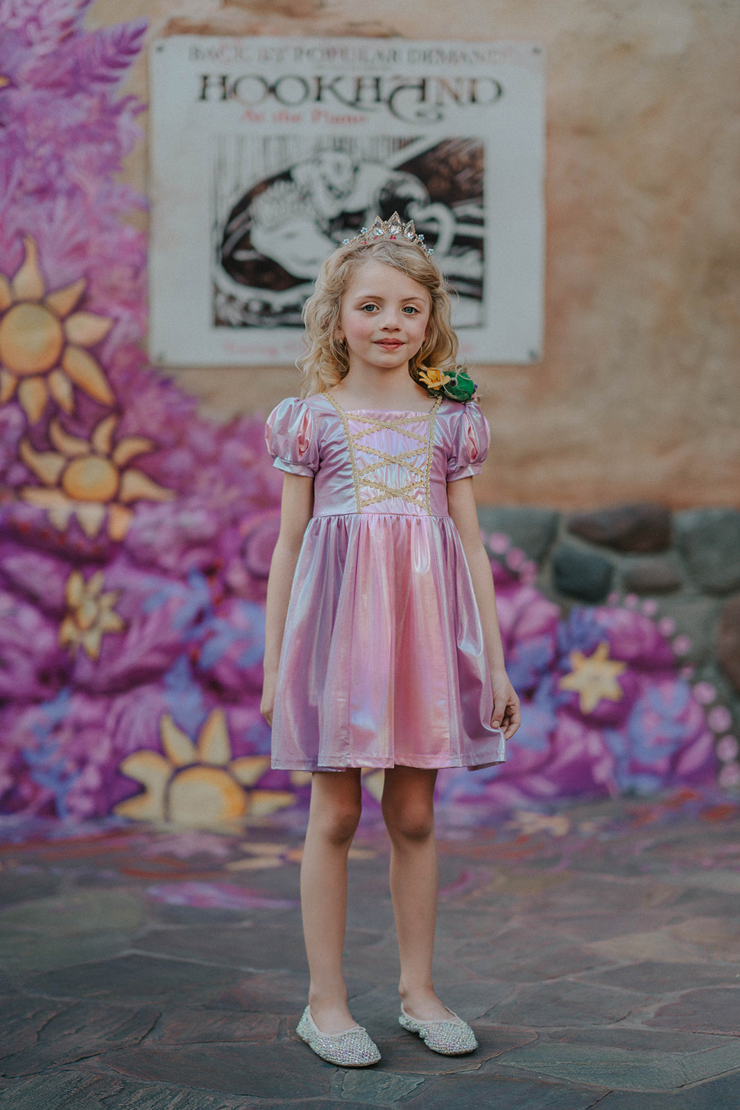 A young girls is wearing a metallic Rapunzel Twirl dress. The pink and purple knee length dress has a criss cross gold ribbon across the bodice. The ballroom puff sleeves add elegance.