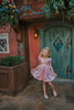 A young girls is wearing a metallic Rapunzel Twirl dress. The pink and purple knee length dress has a criss cross gold ribbon across the bodice. The ballroom puff sleeves add elegance.