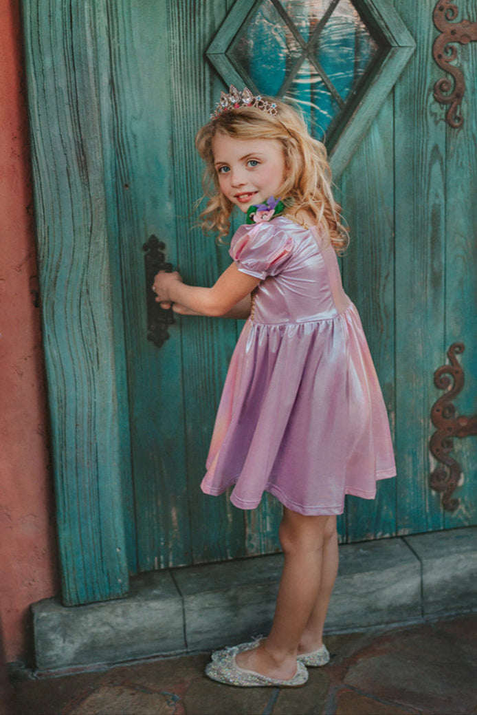 A young girls is wearing a metallic Rapunzel Twirl dress. The pink and purple knee length dress has a criss cross gold ribbon across the bodice. The ballroom puff sleeves add elegance.